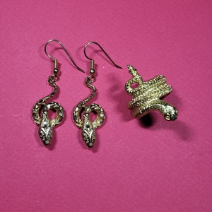 May include: Gold-toned snake jewelry set on a pink background. The set includes a pair of dangle earrings and a ring, all featuring a detailed snake design. The earrings have curved snake bodies, and the ring is a coiled snake.