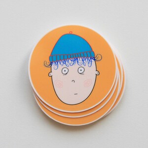 Könnte beinhalten: Ein Stapel runder Aufkleber mit orangefarbenem Hintergrund und einer Cartoon-Illustration einer Person mit einem blauen Hut. Die Person hat blaues Haar und schaut mit einem überraschten Ausdruck geradeaus.