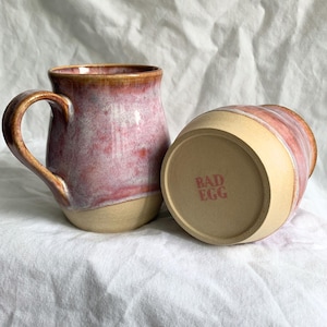 May include: Two ceramic mugs with a pink and beige glaze. One mug is upright, showing the handle and interior. The other mug is upside down, revealing the text "BAD EGG" in red. The mugs are on a white, textured surface.