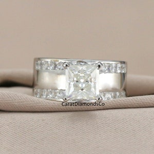 May include: A silver ring featuring a large square-shaped diamond in the centre. The diamond is surrounded by smaller diamonds on either side. The ring is set against a beige fabric background. CaratDiamondsCo is printed on the fabric.
