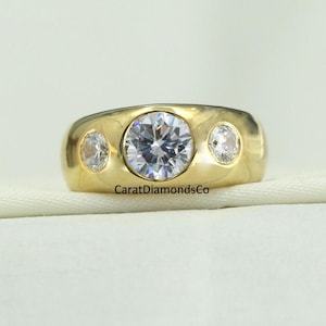 May include: A gold ring with three clear, round gemstones. The center stone is larger than the two side stones. The ring has a smooth, polished finish. The text "CaratDiamondsCo" is visible on the ring.