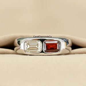 May include: A silver ring featuring two rectangular gemstones: a light yellow and a deep red. The gemstones are set side-by-side in a polished silver band. The ring is displayed against a neutral background.