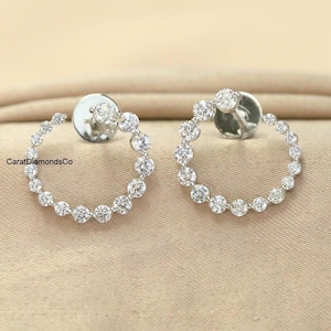 May include: A pair of silver hoop earrings, each adorned with a series of sparkling, round-cut diamonds. The earrings are set against a neutral-toned fabric, highlighting their brilliance. The earrings are a classic jewelry piece.