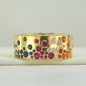 Puede incluir: Un anillo de banda de oro con una colorida variedad de piedras preciosas. El anillo presenta un degradado de colores, incluyendo piedras preciosas azules, rojas, rosas y naranjas. El texto "CaratDiamondsCo" es visible en el anillo.