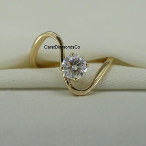 Puede incluir: Un anillo dorado con una sola gema redonda y transparente. La banda del anillo tiene un diseño único y fluido. El texto "CaratDiamondsCo" es visible en el anillo. El anillo está sobre un fondo blanco.