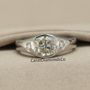 May include: A silver ring with a large, round, clear gemstone in the center and two smaller, round, clear gemstones on either side. The ring is on a light brown fabric background. CaratDiamondsCo.