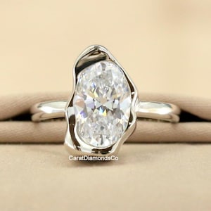 May include: A silver ring with a large, oval-cut diamond. The diamond is set in a unique, organic-shaped bezel. The ring band is smooth and polished. The ring is displayed on a neutral-colored fabric. The text "CaratDiamondsCo" is visible.