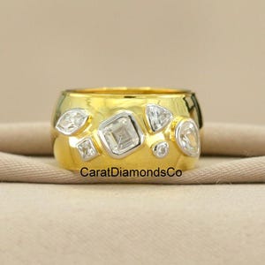 Puede incluir: Un anillo ancho de oro con una serie de piedras preciosas claras de forma irregular. Las piedras preciosas están dispuestas en un patrón alrededor de la banda del anillo. El anillo se muestra sobre una tela de color neutro. El texto "CaratDiamondsCo" es visible en el anillo.