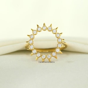 May include: A gold ring with a sunburst design. The ring features a circular arrangement of small, sparkling gemstones, each set within a pointed prong. The ring is displayed on a white cloth.