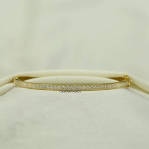 May include: A gold bangle bracelet with a row of small, clear gemstones set along the front. The bracelet is resting on a white surface. The text "CaratDiamondsCo" is visible.