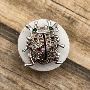 May include: A silver-toned ladybug brooch with red and clear rhinestones and green accents. The brooch is on a white background.