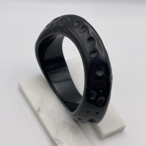 May include: A black plastic bangle bracelet with a textured surface. The bracelet has a circular design with raised dots.