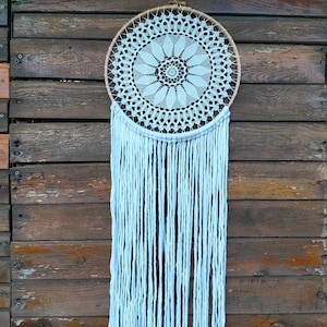 May include: A handmade dreamcatcher with a circular, cream-colored doily center and long, white fringe. The doily features a floral design within a wooden hoop. The dreamcatcher hangs against a weathered, brown wooden background.