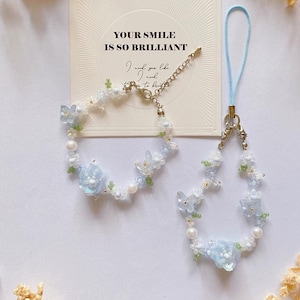 May include: A set of two delicate blue and white floral beaded bracelets with a silver charm. The charm has the text "Your smile is so brilliant" on a white card. The bracelets are on a white background.