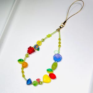 May include: A phone charm featuring a gold-toned cord and a variety of colourful beads. The beads include shapes of red grapes, yellow and blue flowers, a yellow heart, and various green, orange, and blue spherical and leaf-shaped glass accents.