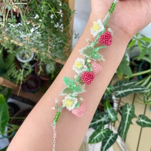 Peut inclure: Un bracelet en perles avec un motif délicat de feuilles vertes, de fleurs blanches et de fraises roses et rouges.