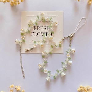 May include: A white and green beaded phone charm with a floral design. The charm features small white flowers and green leaves. The charm is attached to a silver chain and a phone strap.