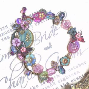 May include: A handcrafted bracelet featuring a floral design with various pastel-colored beads. The bracelet includes pink, purple, and blue flower-shaped beads, along with leaf-shaped beads. A large, round, turquoise-colored bead is also present.