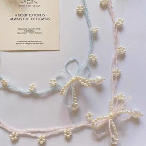 Op de afbeelding: Zachte kralenaccessoires in pastelblauw en roze, versierd met kleine parelachtige bloem- en hartvormige accenten. Het ontwerp bevat een boogachtige structuur. Een kaart met tekst is zichtbaar op de achtergrond.