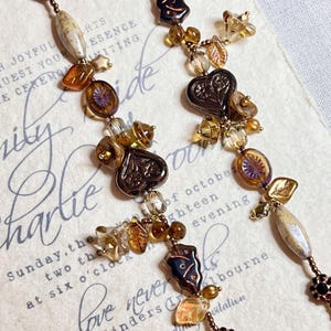 May include: Two long, brown and gold beaded necklaces with heart-shaped charms, leaves, and other decorative elements. The necklaces are hanging on a white background with text.