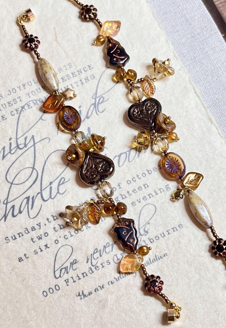 May include: Two long, brown and gold beaded necklaces with heart-shaped charms, leaves, and other decorative elements. The necklaces are hanging on a white background with text.