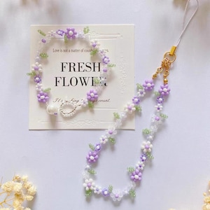 Peut inclure: Un porte-clés de téléphone en perles violettes et blanches avec un accent de perle. Le charme présente de petites fleurs et des feuilles. Le charme est attaché à une chaîne dorée avec un fermoir à homard.
