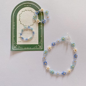 Puede incluir: Un conjunto de dos pulseras de flores de cuentas en azul, blanco y dorado. La pulsera más grande es una cadena delicada de pequeñas flores, mientras que la pulsera más pequeña es un anillo simple.