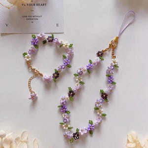 Pode incluir: Um conjunto de joias florais com uma pulseira e uma alça de telefone. A pulseira e a alça são adornadas com pequenas flores de contas em tons de roxo, branco e roxo escuro, com folhas verdes. O conjunto inclui um fecho e uma corrente dourados.