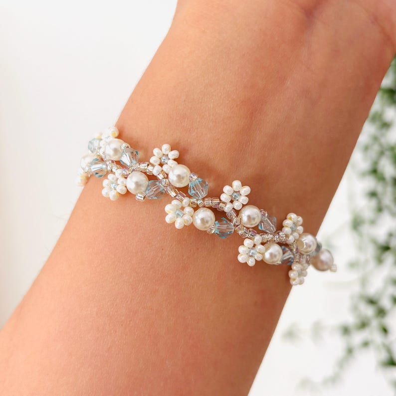 May include: A delicate bracelet featuring a floral design. The bracelet is adorned with white faux pearls, small white flower-shaped beads, and light blue crystal beads. The bracelet is worn on a wrist.