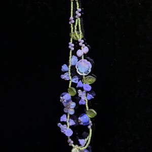 May include: A beaded phone charm featuring two strands of lime green seed beads interspersed with various purple and blue flower-shaped glass beads, translucent light green leaf-shaped beads, and a central iridescent flower-shaped glass bead.