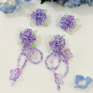 Puede incluir: Un conjunto de pendientes y tachuelas de cuentas hechas a mano con un tema floral. Los pendientes presentan racimos de cuentas moradas y azules con hebras colgantes y acentos de mariposas. Los pendientes y tachuelas tienen acentos de hojas verdes.