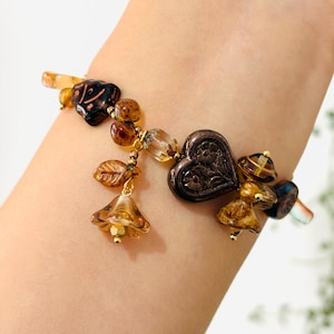 May include: A brown and gold charm bracelet with a heart, leaves, and bells. The bracelet is made of glass beads and metal charms.