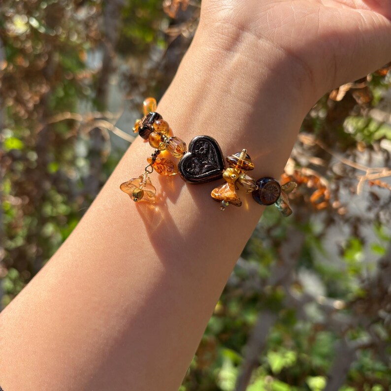 May include: A brown and gold beaded bracelet with a heart-shaped charm. The bracelet is made of glass beads and has a delicate, feminine look.