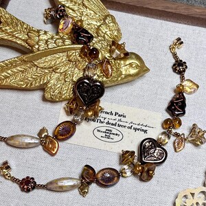 May include: A brown and gold necklace with heart-shaped charms, leaves, and other decorative elements. The necklace is on a fabric surface with a small card that reads "French Paris try and have puthebur you The dead tree of spring Wooden jewelry box".