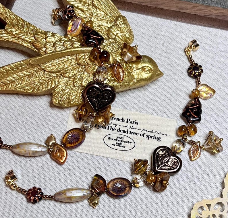 May include: A brown and gold necklace with heart-shaped charms, leaves, and other decorative elements. The necklace is on a fabric surface with a small card that reads "French Paris try and have puthebur you The dead tree of spring Wooden jewelry box".