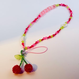 May include: A beaded phone charm with a cherry and flower design. The charm features red, pink, and green beads, with the word "VICTORY" spelled out in pink beads. The charm has a red cherry pendant.