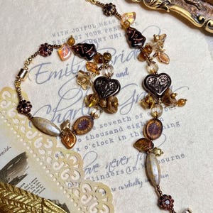 Puede incluir: Un collar de cuentas marrones y doradas con dijes en forma de corazón, hojas y flores. El collar está sobre un fondo blanco con texto que dice "Con corazones alegres, les solicitamos su presencia en la ceremonia que une a Emily y Charles."