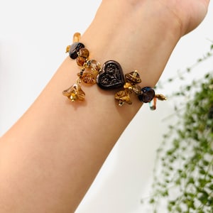 May include: A brown and gold beaded bracelet with a heart-shaped charm. The bracelet features a variety of glass beads in shades of brown, gold, and amber. The heart charm is made of metal and has a detailed design.