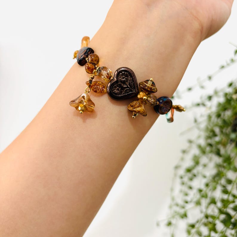May include: A brown and gold beaded bracelet with a heart-shaped charm. The bracelet features a variety of glass beads in shades of brown, gold, and amber. The heart charm is made of metal and has a detailed design.