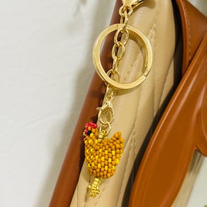 May include: A gold-colored keychain with a beaded chicken charm. The chicken is yellow, orange, and red, with a gold-colored beak and feet. The keychain is attached to a gold-colored ring and clasp.