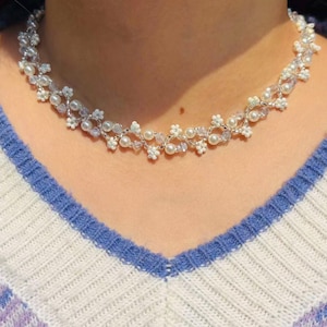 May include: A delicate necklace featuring a floral design with white pearls and small white flower accents. The necklace is made of silver-colored metal and is worn against a white, blue, and purple striped sweater.