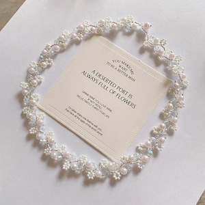May include: A delicate, handcrafted floral headpiece with white pearls and iridescent crystals. The headpiece surrounds a card with the text "A Deserted Port Is Always Full of Flowers."