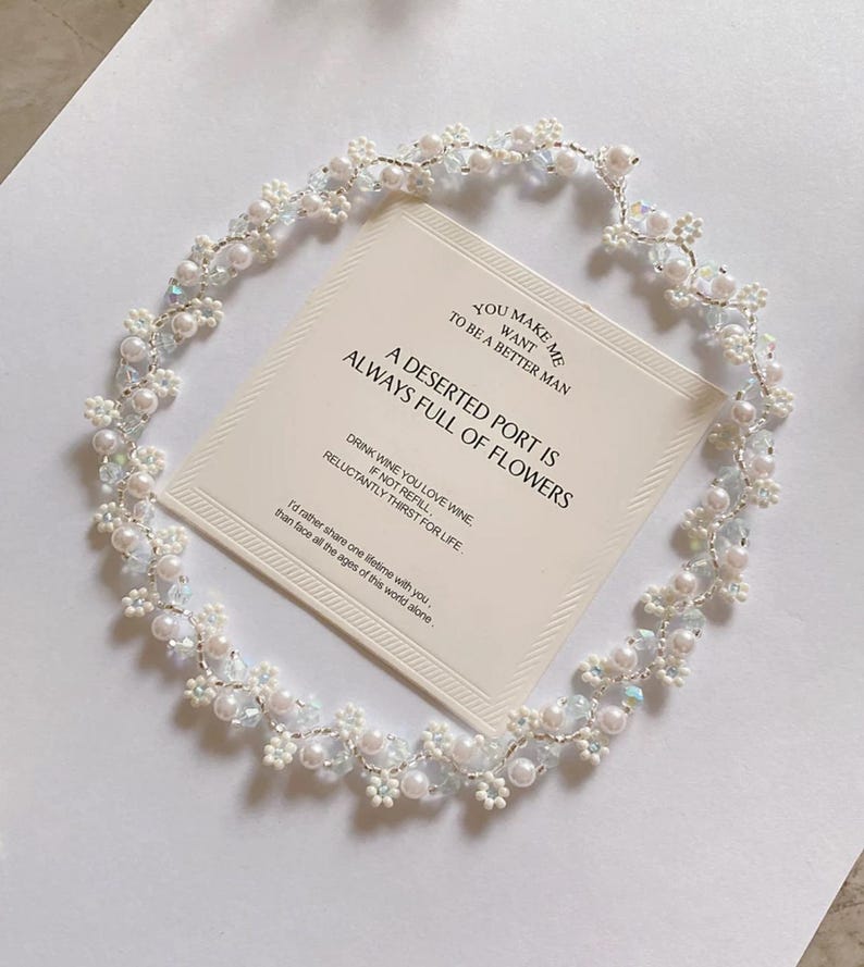 May include: A delicate, handcrafted floral headpiece with white pearls and iridescent crystals. The headpiece surrounds a card with the text "A Deserted Port Is Always Full of Flowers."