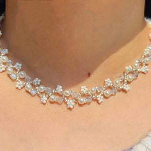 May include: A delicate necklace featuring a floral design with white pearls, clear crystals, and small white flower accents. The necklace is made with silver-colored wire and is designed to sit around the neck.