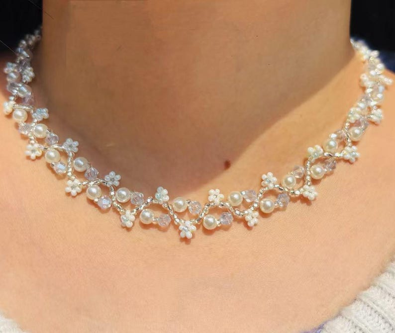 May include: A delicate necklace featuring a floral design with white pearls, clear crystals, and small white flower accents. The necklace is made with silver-colored wire and is designed to sit around the neck.