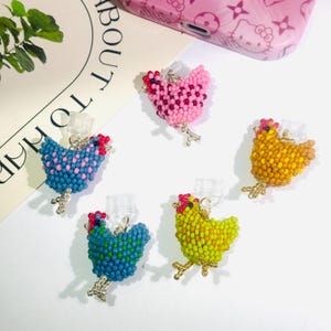 Peut inclure: Plusieurs breloques en forme de poulet, faites de perles de différentes couleurs : rose, bleu, jaune et vert. Chaque breloque a une crête rouge et des pattes argentées. Ces breloques sont conçues pour être attachées à un téléphone ou autre appareil.