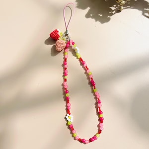 May include: A pink, red, and green beaded phone charm with a strawberry charm and a white flower charm.
