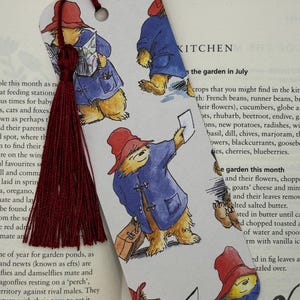May include: A rectangular paper bookmark featuring a repeating illustration of a bear wearing a red hat and blue coat, with a dark red tassel attached through a hole at the top. The bear is depicted in various poses, including reading a map and holding a suitcase.