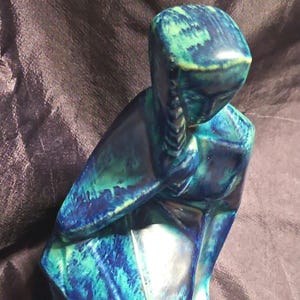 Könnte beinhalten: Eine Keramikskulptur einer Figur mit geflochtener Frisur, versehen mit einer glänzenden Glasur in gemischten Schattierungen von tiefem Blau, Türkis und Petrol. Die Figur ist in sitzender oder kniender Haltung mit einem erhobenen Arm dargestellt.