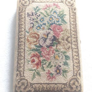 Könnte beinhalten: Eine beige Nadelmalerei-Box mit einem Blumendesign. Das Design zeigt einen Strauß aus rosa, roten, blauen und gelben Blumen mit grünen Blättern. Die Box ist von einem braunen Rahmen umgeben.